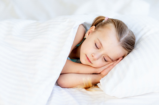 a girl child sleeps on a bed at home on a white cotton bed and smiles sweetly in her sleep with her hands folded under her cheeks