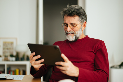 Smiling mature man reading business report on his tablet while sitting at home office desk with documents.