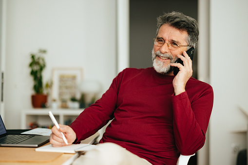 Happy senior business man speaking on the phone while sitting at home office desk with laptop computer and writing notes in a notebook.