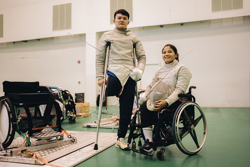 A young fencer wearing prosthetic legs and a female fencer in a wheelchair warm up in the gym before the match.
