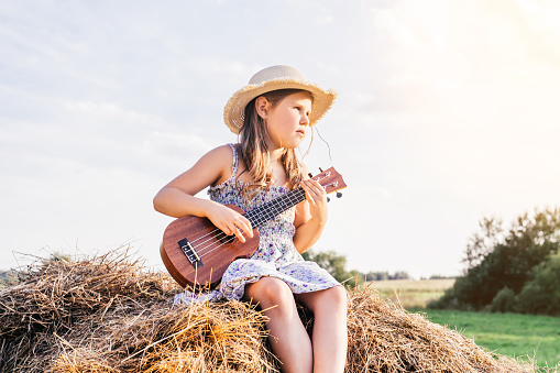 Pretty little girl in panama hat sit at top of haystack looking away and play ukulele. Carefree child resting on hayrick. Outdoor walking. Beautiful sunny sky. Summer vacation, countryside lifestyle.