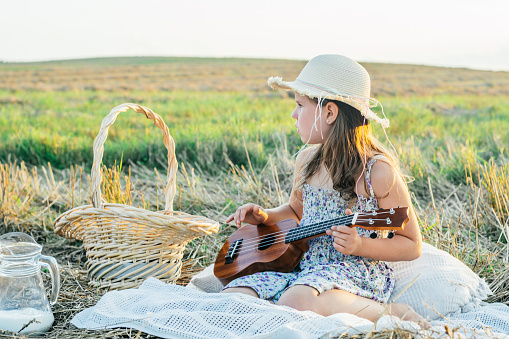 Little girl play ukulele sitting on blanket on grassy field. Portrait of child in panama hat. Outdoor picnic with food basket. Music instrument, hobby. Summer vacation, countryside lifestyle.