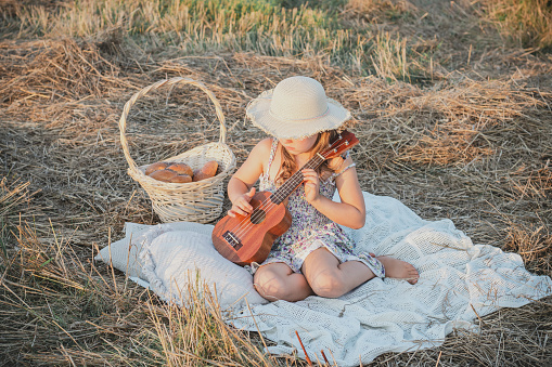 Little girl play ukulele sitting on blanket on field among dry cut grass. Portrait of child in panama hat. Outdoor walking and picnic. Music instrument, hobby. Summer vacation, countryside lifestyle.