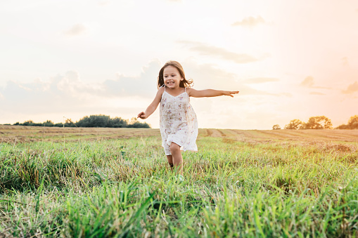 Happy little girl joyfully running across field. Carefree child on grassy meadow. Outdoor walking. Beautiful sunny sky. Summer vacation, countryside lifestyle. Picturesque landscape.