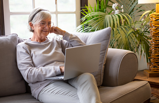 Happy 70s white-haired woman sitting on couch in living room, using laptop watching movie. Older generation and wireless technology users concept