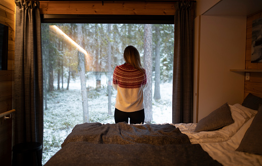 Back view of a woman in a sweater standing in a cottage looking out the window