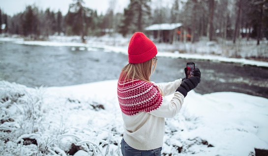 A woman wearing winter gloves, a cap and a sweater takes a selfie while standing by the river