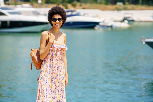 Happy black woman in dress and sunglasses with bag, on shoulder smiling and looking at camera on blurred background on sea and boats on summer day in harbor