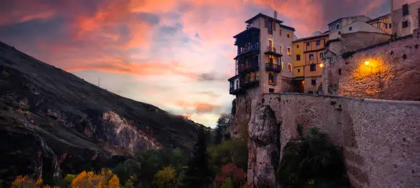 sunset with a view of the Hanging Houses of Cuenca, Spain, with a rock, a sky with blue, orange and pink clouds, and a dark landscape of the city. Tourist postcard or wallpaper from Castilla