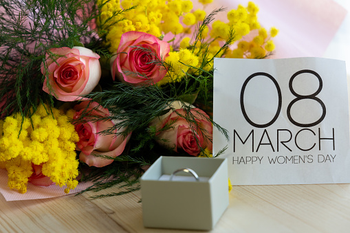 Close-up view of a bouquet of roses and mimosa flowers, wedding ring on a ring box and 8 march sign on a wooden table