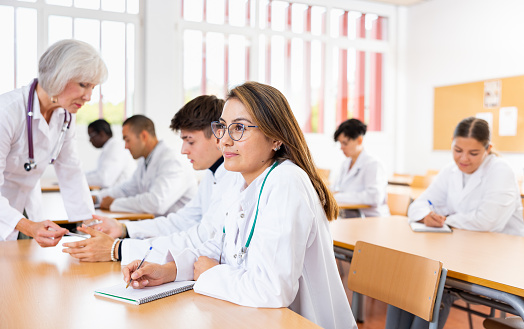 Interested young latin american woman in white coat listening to lecture and taking notes in classroom during professional medical training