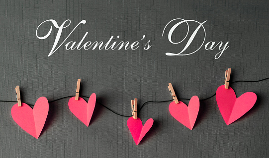 Pink paper cut hearts hanging on a clothesline with wooden clothespins. VALENTINE'S DAY text is in the center. Can be used as a design for Valentine's day holiday greeting cards or posters.