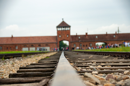 A low angle focused on the railway track at the entrance of the Auschwitz concentration camp. It's a cloudy day in the summer with the brick wall entrance and guard Tower in the background