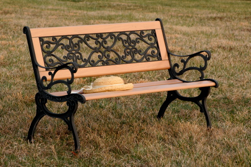 Classic straw hat with ivory grosgrain ribbon eft behind on wrought iron and wooden park bench in the grass