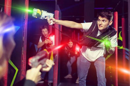 Emotional young man with laser pistol playing laser tag with friends on dark labyrinth