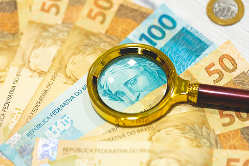 Brazilian Real banknotes on a wooden table with a golden magnifying glass in the composition. Brazilian economy, finance and inflation.