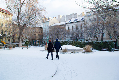 Budapest, Hungary - January 22, 2022 - Young Heterosexual Couple Walking in Snowy Garden in Budapest, Rear View, Károlyi Garden Downtown Budapest Cityscape, Public Park After Morning Snowfall as Increasingly Rare Natural Phenomenon in the Hungarian Capital in the 21st Century, Hungarian National Museum Neighborhood, in Inner City 5th District on Day of the Hungarian Culture