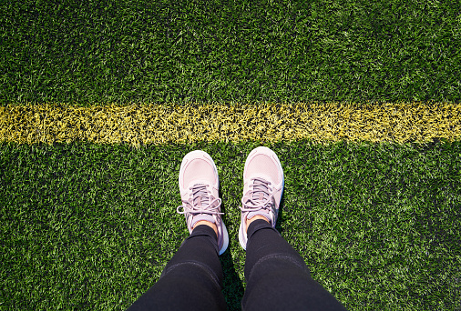 Legs of a woman standing on a green sports playground in front of a yellow line, top view