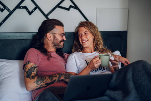 pareja romántica acostada en la cama de casa por la mañana y usando una computadora portátil - hotel newspaper coffee reading fotografías e imágenes de stock