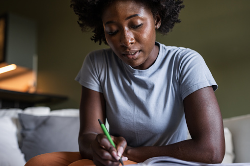 A young African-American woman is doing home budgeting. She is sitting on the couch and writing a list of her bills.