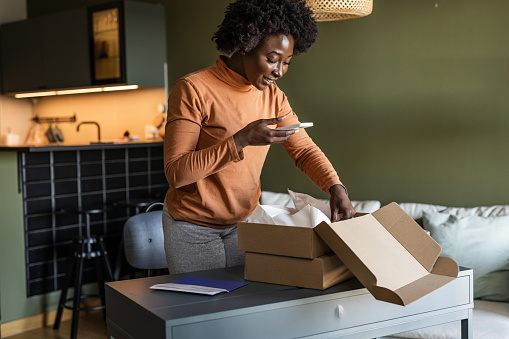 A smiling young African-American woman is unpacking a carton box, excited to see her online purchase. She is taking pictures of her delivery for social media.
