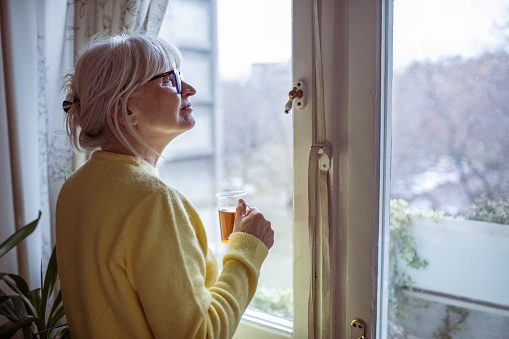 Mature woman in a yellow sweater drinking tea by a window, looking outside