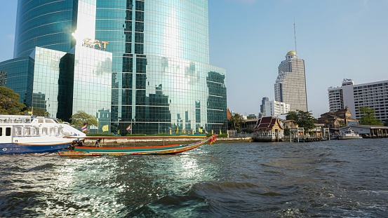 Bangkok, Thailand-February 6, 2019: Chao Phraya riverside of the Thai capital with a view of tall modern glass skyscrapers in the background and landmarks of the city next to the river where people can take a water taxi to explore the city. Worldwide famous tourist destination.
