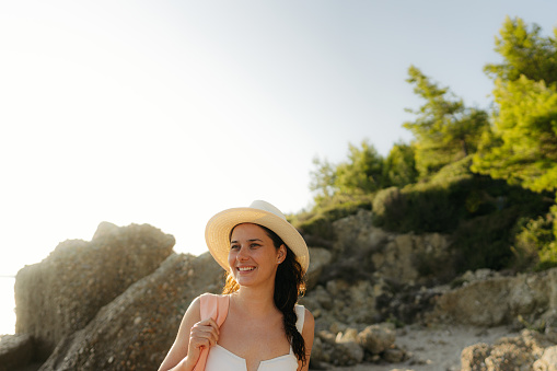 Photo of a smiling young woman, wearing her hat and a beach bag, ready to find some cozy corner at a lonely beach