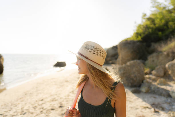 Photo of a smiling young woman, wearing her hat and a beach bag, ready to find some cozy corner at a lonely beach