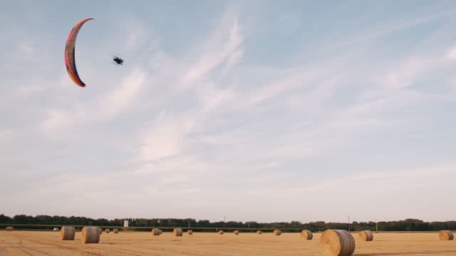 Slow motion handheld shot of a paraglider flying over a harvested agricultural field full of ton bales