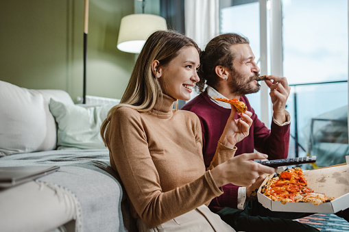 Caucasian couple enjoying free time at home. They are watching TV in the living room while eating pizza