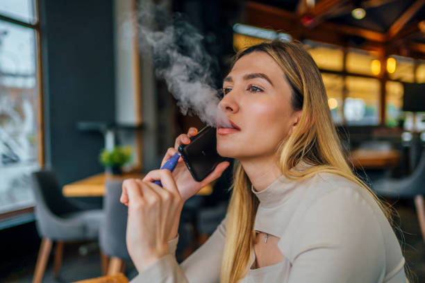 Woman smokes disposable vape cigarette while talking on smart phone stock photo