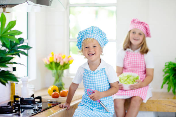 Kids cook in white kitchen. Children cooking at home. Little boy and girl in chef hat and apron cut vegetables for salad. Healthy nutrition for young child. Kid helping to prepare dinner.