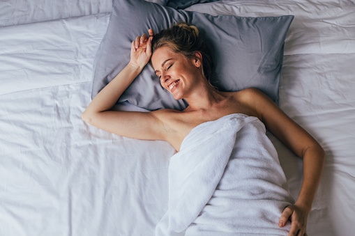 A from above view of a smiling Caucasian female wrapped in a towel lying on the bed with her eyes closed after having a shower.