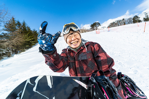 A senior Japanese woman wearing snowboarding clothing and holding her snowboard on a ski run holding up the 