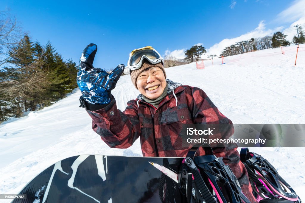 A cheerful young at heart senior snowboarder woman with her snowboard giving a "devil horns" hand gesture A senior Japanese woman wearing snowboarding clothing and holding her snowboard on a ski run holding up the "devil horns" hand gesture such as a young person might do. Happiness Stock Photo