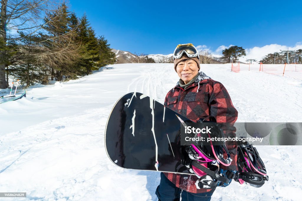 A young at heart cheerful senior snowboarder woman with her snowboard A senior Japanese woman wearing snowboarding clothing and holding her snowboard on a ski run. Happiness Stock Photo