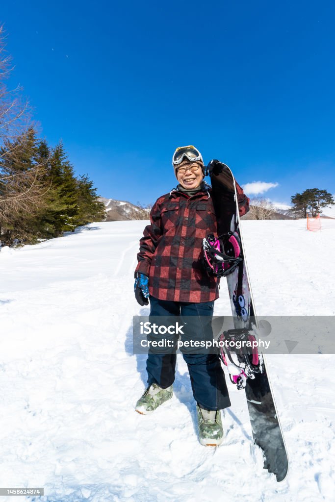 A young at heart cheerful senior snowboarder woman with her snowboard A senior Japanese woman wearing snowboarding clothing and holding her snowboard on a ski run. 60-64 Years Stock Photo