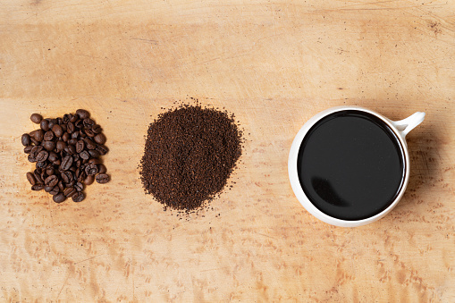 Whole coffee beans, ground coffee, and a cup of brewed black coffee on a wood table, top view