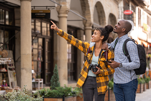 Young african couple of tourists with smartphone looking for destination and taking pictures on camera while walking on the street in an old town in Europe. Woman pointing interesting place
