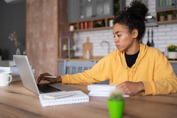 istock An african-american young woman is using laptop computer for remote work or studying online, she takes notes watching webinars or classes at kitchen table at home 1458681170