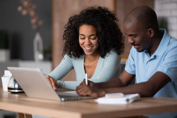 istock Modern young African American couple watching online learning classes, sitting in the kitchen, taking    notes and discussing, e-learning concept 1458681173