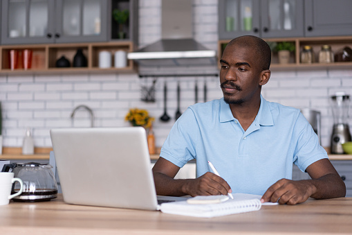 Online Education. Serious african man studying and working online, watching webinar on a laptop and taking notes. African man on a distance learning, sitting at kitchen table at home