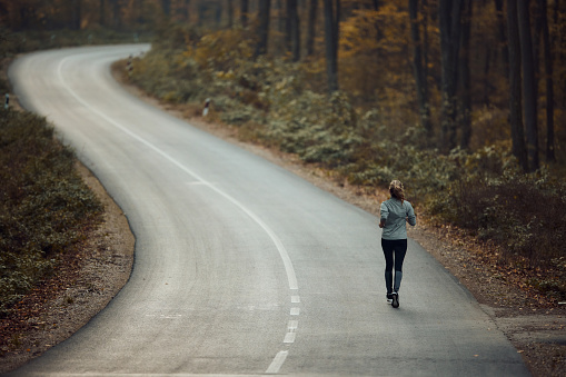 Back view of athletic woman running on the road during autumn day in the forest. Copy space.
