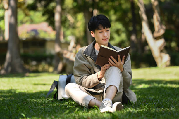 istock Happ young asian man reading interesting book while while sitting on green grass at sunny beautiful day. 1458693105