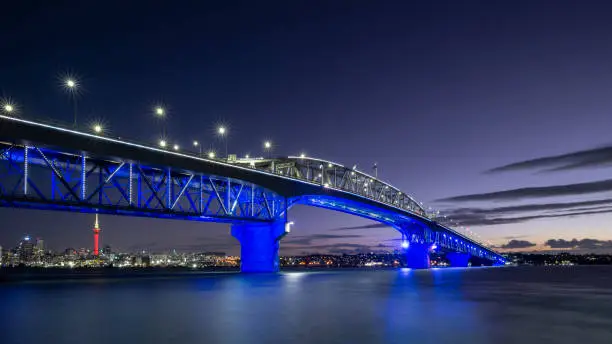 Auckland Harbour Bridge glowing in blue. Skytower in gold and red. Auckland, New Zealand