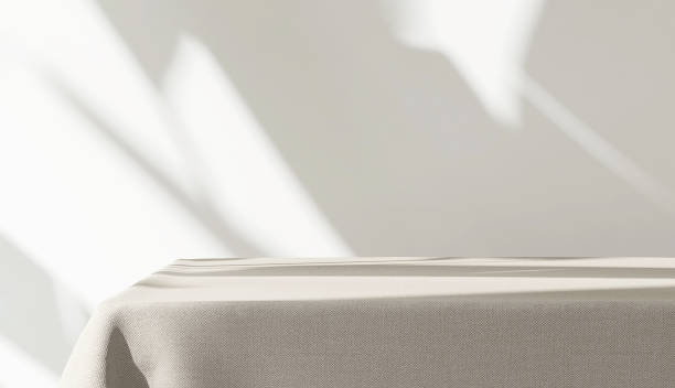 istock Table counter with beige brown tablecloth drape in sunlight, tropical leaf shadow on blank white wall 1458700242