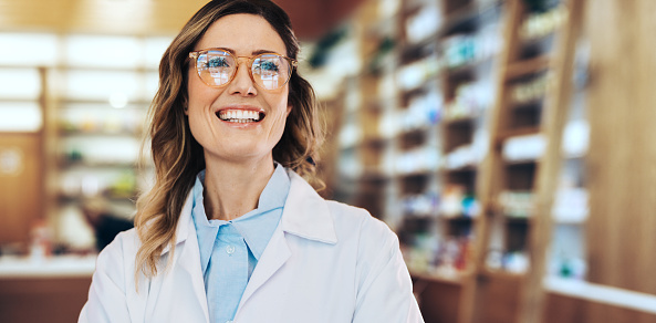 Healthcare worker standing in a pharmacy in a white coat. Woman working in a drug store. Female pharmacist smiling.