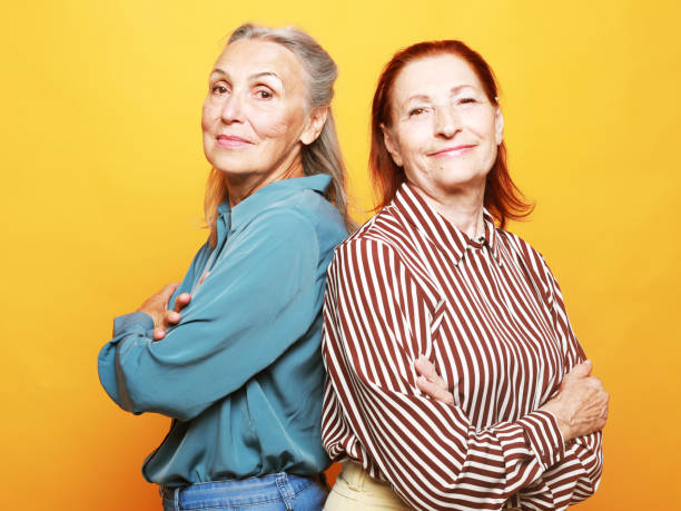 Two elderly women friends with crossed arms on yellow background. Lifestyle concept. stock photo