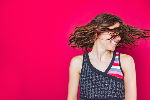 Portrait of young woman in sleeveless top tossing her hair, smiling happily and turning head to her left on vivid magenta background